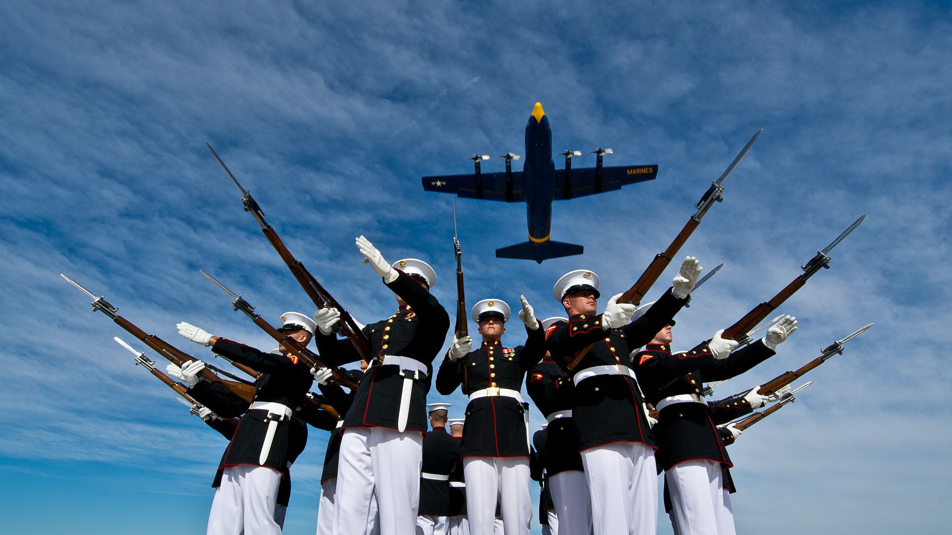 Marines stand at attention and salute with rifles while blue angels c-130 flies overhead during daytime